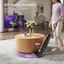 A woman in a lavender top and cream pants vacuums a light gray rug in a modern living room. The upright vacuum is shown from the side with a 90-degree angle graphic, and a floating inset highlights the front propulsion feature.